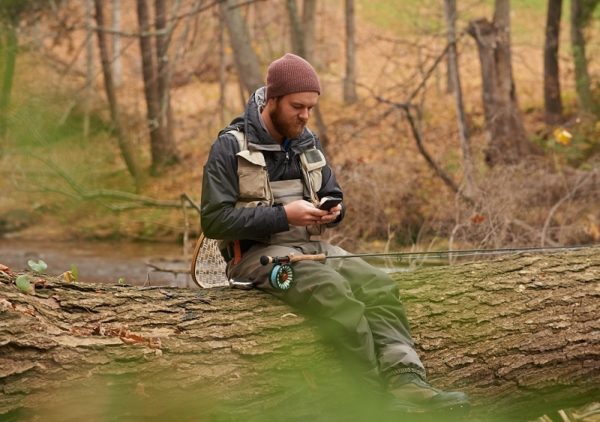 Fisherman sitting on a fallen tree trunk looking at his phone